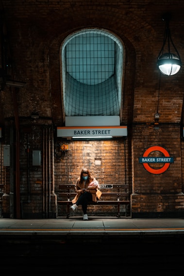 A person wearing a mask is sitting on a bench at Baker Street underground station. The brick walls and dim lighting create a warm, moody atmosphere. The iconic London Underground sign is visible, and the person appears to be engrossed in their phone, holding a bouquet of flowers.