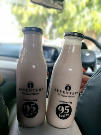 Two Keventers milkshake bottles are positioned in a car interior. The bottles are labeled with the brand name and a celebration marking 95 years. The surrounding environment is partially visible with car seats and a blurred view outside showing greenery.