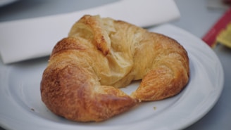A buttery croissant resting on a clean white plate with a folded white napkin nearby. The croissant has a golden-brown, flaky texture, indicating it is freshly baked. In the background, part of a red and yellow packaging is visible, slightly blurred.