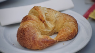 A buttery croissant resting on a clean white plate with a folded white napkin nearby. The croissant has a golden-brown, flaky texture, indicating it is freshly baked. In the background, part of a red and yellow packaging is visible, slightly blurred.