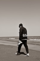 A lone individual walking barefoot on a sandy beach while holding a pair of sneakers in one hand. The person is dressed in black clothing that features the names of various cities. The ocean waves gently approach the shore, and the sky is clear.