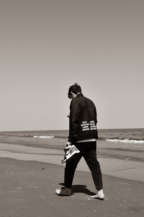 A lone individual walking barefoot on a sandy beach while holding a pair of sneakers in one hand. The person is dressed in black clothing that features the names of various cities. The ocean waves gently approach the shore, and the sky is clear.
