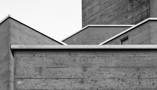 A black and white photograph of a modern architectural structure featuring geometric shapes and clean lines. The focus is on the intersecting angles and surfaces of the building, with a stark contrast between the light sky and the textured concrete walls.