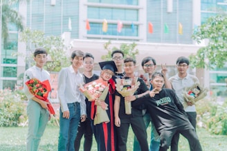 A group of eight young people are posing happily outdoors. One person is wearing a graduation cap and gown, holding a bouquet of flowers, indicating a graduation celebration. Several others also hold bouquets and gifts. The background features a modern building with colorful flags and greenery, suggesting a festive occasion.