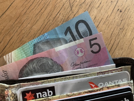 A close-up view of a wallet containing Australian currency notes, specifically a five and ten dollar bill, alongside several cards including ones marked 'nab' and 'Qantas'. The surface beneath the wallet is a wooden table with visible grains.