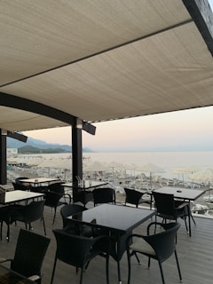 An outdoor patio of a beachside restaurant features wooden tables and wicker chairs under a large canopy. In the background, multiple sun loungers and umbrellas line the sandy beach, with calm sea waters and distant mountains visible under a clear sky.
