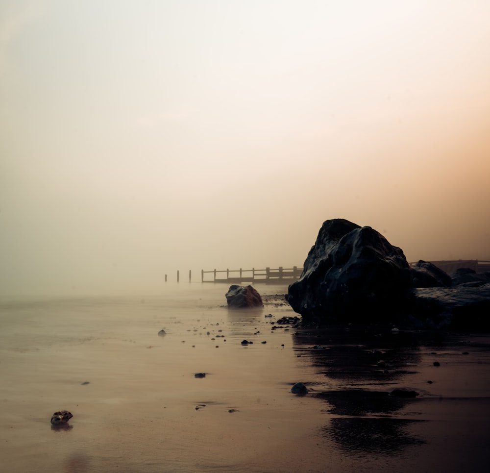 a foggy beach with rocks and a bridge in the distance