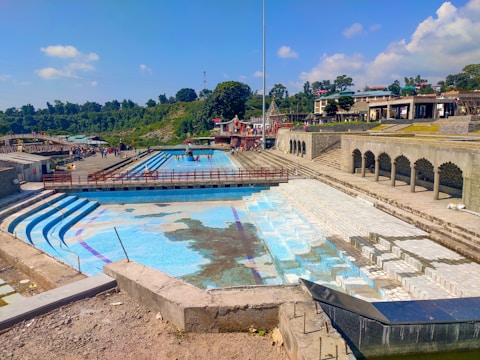 A large stepped water feature with empty and partially tiled pools is surrounded by concrete pathways. There are structures with arched openings along one side, and lush greenery with trees and shrubs on the other. People are seen walking and gathering in the open areas under a clear blue sky with a few scattered clouds.