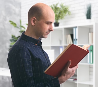 A man is standing indoors, holding and reading a large red book. He is wearing a dark, checkered shirt and is positioned in front of a white bookshelf filled with various books and decorative items. Green plants are placed at the top of the shelf, adding a touch of nature to the setting.
