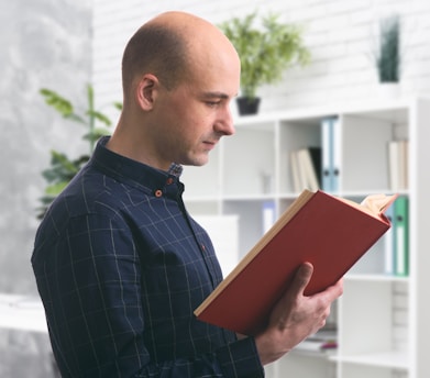 A man is standing indoors, holding and reading a large red book. He is wearing a dark, checkered shirt and is positioned in front of a white bookshelf filled with various books and decorative items. Green plants are placed at the top of the shelf, adding a touch of nature to the setting.