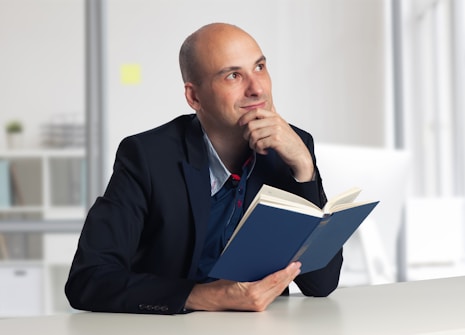 A man in a dark suit is seated at a lightly colored table, holding a blue book and gazing thoughtfully upwards. The background is softly blurred, suggesting an office or library setting with shelves and a window.