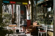A quaint outdoor seating area of a cafe with a vintage vibe, featuring a piano and mismatched wooden chairs. Potted plants and hanging greenery line the window ledge, while sunlight filters through, casting soft shadows. The storefront has ornate details and a sign indicating 'espresso.'