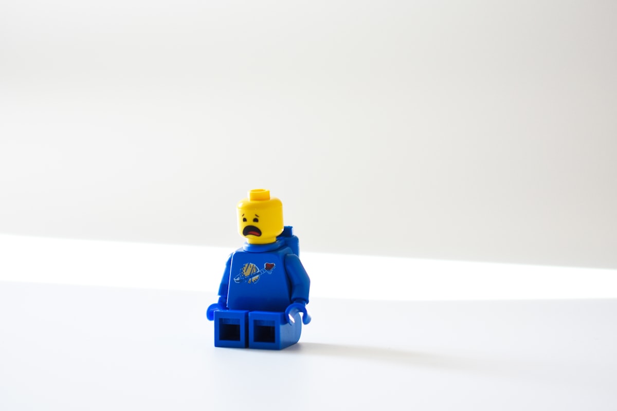 A lego figure wearing all blue it a sitting pose in a white space.  It has a yellow head and is upset which is shown by a large, open, downturned mouth.