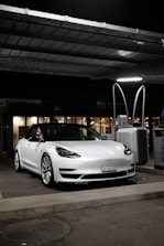 A sleek, white Tesla car, parked under a canopy at a gas station or charging point during nighttime. The car is positioned near a charging station, and the surroundings are dimly lit with a few lights visible in the background.