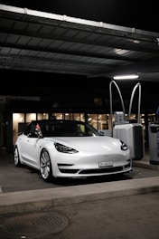 A sleek, white Tesla car, parked under a canopy at a gas station or charging point during nighttime. The car is positioned near a charging station, and the surroundings are dimly lit with a few lights visible in the background.