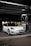 A sleek, white Tesla car, parked under a canopy at a gas station or charging point during nighttime. The car is positioned near a charging station, and the surroundings are dimly lit with a few lights visible in the background.