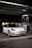 A sleek, white Tesla car, parked under a canopy at a gas station or charging point during nighttime. The car is positioned near a charging station, and the surroundings are dimly lit with a few lights visible in the background.
