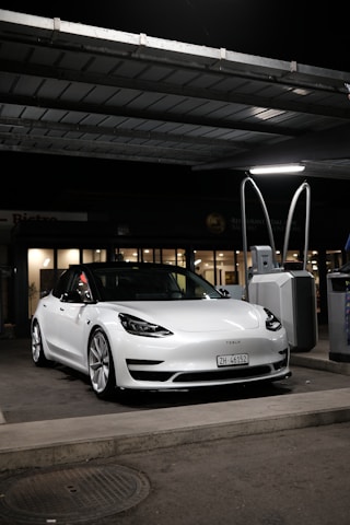 A sleek, white Tesla car, parked under a canopy at a gas station or charging point during nighttime. The car is positioned near a charging station, and the surroundings are dimly lit with a few lights visible in the background.