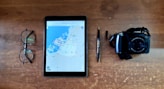 A wooden table surface with a pair of round glasses, a tablet displaying a map, a black pen, and a digital camera. The objects are neatly arranged in a row, suggesting a theme of travel or preparation.