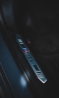 A close-up of a car door sill featuring a metallic plate engraved with the logo 'BMW' and the M division stripes in blue, purple, and red. The surroundings are dark and sleek, suggesting a luxurious vehicle interior.
