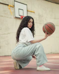 woman in white long sleeve shirt and gray pants sitting on brown wooden floor holding basketball