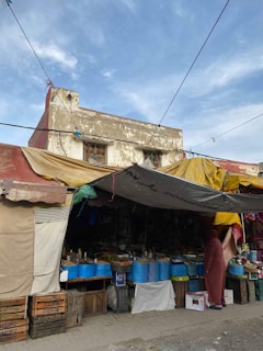 A street market scene with a variety of goods displayed under a canopy. Several large blue buckets are prominently displayed on tables, with various packaged products. The market stall is housed under a makeshift canopy made from different colored tarps, adding to the eclectic and vibrant atmosphere. A person dressed in a long robe stands to one side, possibly browsing the items for sale. The background features an old, weathered building with a hint of the sky above.