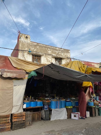 A street market scene with a variety of goods displayed under a canopy. Several large blue buckets are prominently displayed on tables, with various packaged products. The market stall is housed under a makeshift canopy made from different colored tarps, adding to the eclectic and vibrant atmosphere. A person dressed in a long robe stands to one side, possibly browsing the items for sale. The background features an old, weathered building with a hint of the sky above.