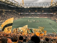 A large, packed stadium during a soccer match with fans waving yellow and black flags, creating a lively and vibrant atmosphere. The crowd is dense, with many people visibly engaged and cheering. Players can be seen on the field, along with some officials and staff.