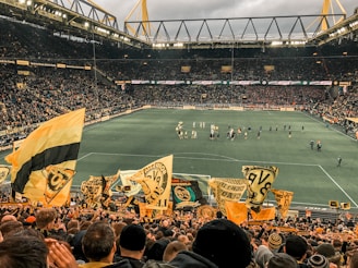 A large, packed stadium during a soccer match with fans waving yellow and black flags, creating a lively and vibrant atmosphere. The crowd is dense, with many people visibly engaged and cheering. Players can be seen on the field, along with some officials and staff.