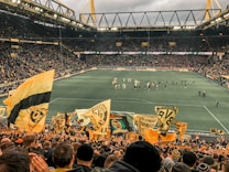 A large, packed stadium during a soccer match with fans waving yellow and black flags, creating a lively and vibrant atmosphere. The crowd is dense, with many people visibly engaged and cheering. Players can be seen on the field, along with some officials and staff.