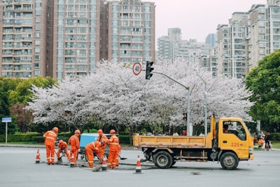 A group of construction workers in bright orange uniforms engage in road work on a city street. A yellow construction truck is parked nearby. The scene is set against a backdrop of blooming cherry blossom trees and modern apartment buildings.