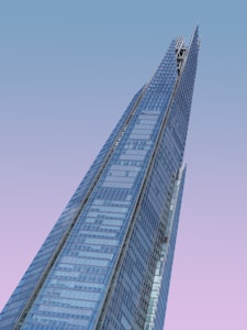 A modern skyscraper with a sleek, angular design and a facade made primarily of glass panels. The background features a gradient sky transitioning from light blue to pink, suggesting a sunrise or sunset. The building has a distinct, pointed top that adds to its futuristic appearance.