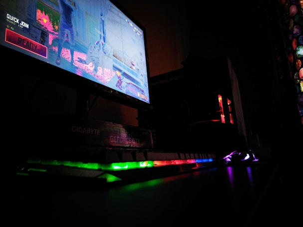 A dimly lit gaming setup is highlighted with a computer monitor displaying a game screen. The surrounding area is dark, with RGB lighting from a keyboard and PC tower creating vibrant reflections on the desk.