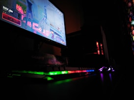 A dimly lit gaming setup is highlighted with a computer monitor displaying a game screen. The surrounding area is dark, with RGB lighting from a keyboard and PC tower creating vibrant reflections on the desk.
