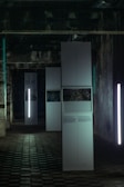 A dimly lit industrial room features tall, white rectangular panels. Some panels display photos and text, while vertical lights provide illumination. The ceiling is marked by exposed pipes and the floor is covered in a grid-like pattern.