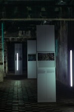 A dimly lit industrial room features tall, white rectangular panels. Some panels display photos and text, while vertical lights provide illumination. The ceiling is marked by exposed pipes and the floor is covered in a grid-like pattern.
