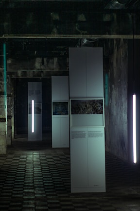 A dimly lit industrial room features tall, white rectangular panels. Some panels display photos and text, while vertical lights provide illumination. The ceiling is marked by exposed pipes and the floor is covered in a grid-like pattern.