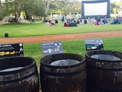 A grassy outdoor area features a large inflatable movie screen with people sitting on blankets to watch an open-air film. In the foreground, three wooden trash bins are labeled for landfill, compostable, and recyclable waste, promoting waste separation and environmental awareness. The environment is surrounded by trees and a relaxed atmosphere is evident.