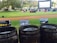 A grassy outdoor area features a large inflatable movie screen with people sitting on blankets to watch an open-air film. In the foreground, three wooden trash bins are labeled for landfill, compostable, and recyclable waste, promoting waste separation and environmental awareness. The environment is surrounded by trees and a relaxed atmosphere is evident.