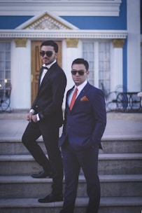 Two men in formal suits stand confidently on steps in front of an elegant building with classical architecture. Both wear sunglasses and have their hands in their pockets, exuding a sense of style and sophistication. The building features large windows and decorative elements with blue and gold accents.