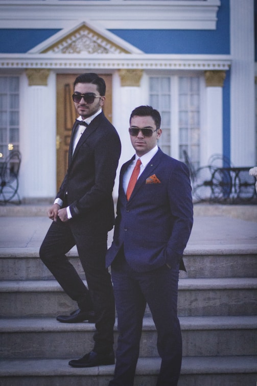 Two men in formal suits stand confidently on steps in front of an elegant building with classical architecture. Both wear sunglasses and have their hands in their pockets, exuding a sense of style and sophistication. The building features large windows and decorative elements with blue and gold accents.