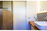 A modern bathroom corner with a white rectangular sink on a wooden countertop. The wall has an artistic geometric patterned backsplash in blue and white. A small potted plant with purple flowers adds a touch of color. The adjacent wall houses a light switch and temperature control panel. The room has a minimalistic design with a natural stone tiled shower area.