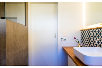A modern bathroom corner with a white rectangular sink on a wooden countertop. The wall has an artistic geometric patterned backsplash in blue and white. A small potted plant with purple flowers adds a touch of color. The adjacent wall houses a light switch and temperature control panel. The room has a minimalistic design with a natural stone tiled shower area.