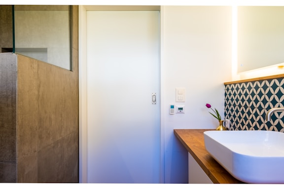 A modern bathroom corner with a white rectangular sink on a wooden countertop. The wall has an artistic geometric patterned backsplash in blue and white. A small potted plant with purple flowers adds a touch of color. The adjacent wall houses a light switch and temperature control panel. The room has a minimalistic design with a natural stone tiled shower area.