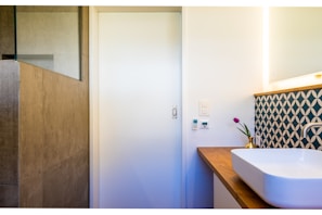 A modern bathroom corner with a white rectangular sink on a wooden countertop. The wall has an artistic geometric patterned backsplash in blue and white. A small potted plant with purple flowers adds a touch of color. The adjacent wall houses a light switch and temperature control panel. The room has a minimalistic design with a natural stone tiled shower area.