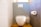 A modern bathroom features a white toilet positioned against a decorative wall with a geometric blue and white tile pattern. To the right, a roll of toilet paper is mounted on a gray wall. The floor is covered in wooden tiles, and there is a metallic toilet brush holder beside the toilet.