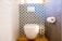 A modern bathroom features a white toilet positioned against a decorative wall with a geometric blue and white tile pattern. To the right, a roll of toilet paper is mounted on a gray wall. The floor is covered in wooden tiles, and there is a metallic toilet brush holder beside the toilet.