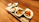 A rectangular ceramic plate holds four small, white, boat-shaped bowls filled with a Japanese dish. Each bowl contains a different type of sushi or sashimi, garnished with green onion and drizzled with a dark sauce. The plate is on a wooden surface, and there is a green leaf placed on the plate under the bowls, providing a contrast with the white bowls and the colorful food.