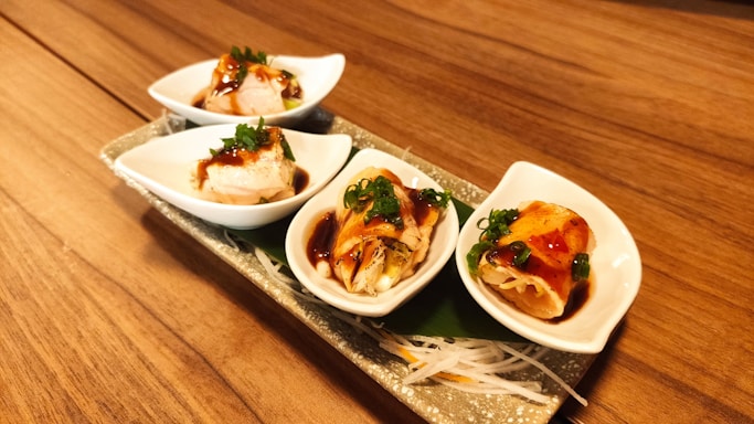A rectangular ceramic plate holds four small, white, boat-shaped bowls filled with a Japanese dish. Each bowl contains a different type of sushi or sashimi, garnished with green onion and drizzled with a dark sauce. The plate is on a wooden surface, and there is a green leaf placed on the plate under the bowls, providing a contrast with the white bowls and the colorful food.