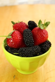 A bright green bowl filled with fresh strawberries and mulberries is placed on a wooden surface. The strawberries are vibrant red, with green leaves still attached, contrasting with the deep purple-black mulberries.